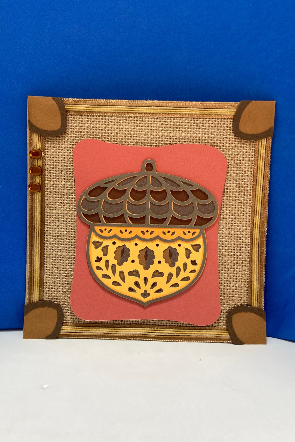 Handcrafted autumn decorative square with a layered mandala-style acorn and burlap-style paper background.