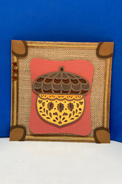 Handcrafted autumn decorative square with a layered mandala-style acorn and burlap-style paper background.