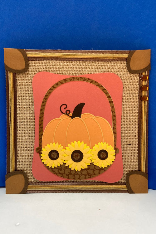 Autumn decorative square featuring a layered pumpkin in a basket with yellow sunflowers on burlap-style paper.